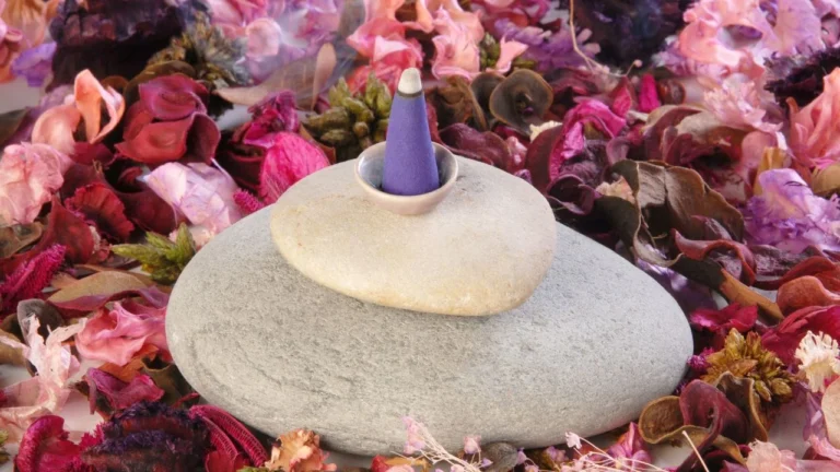 Fioletowy stożek kadzidła umieszczony na kamieniu, otoczony suszonymi kwiatami w odcieniach różu i czerwieni.